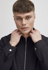 Svart zippered jakke med spiss krage, knappelukking på mansjettene og teksturert innerfôr. Modellen holder kragen med hendene. Ren bakgrunn.