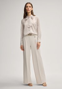 Donna con pantaloni beige a gamba larga, camicetta a maniche lunghe con fantasia e fiocco, cintura dorata e sandali aperti in piedi davanti a uno sfondo semplice.