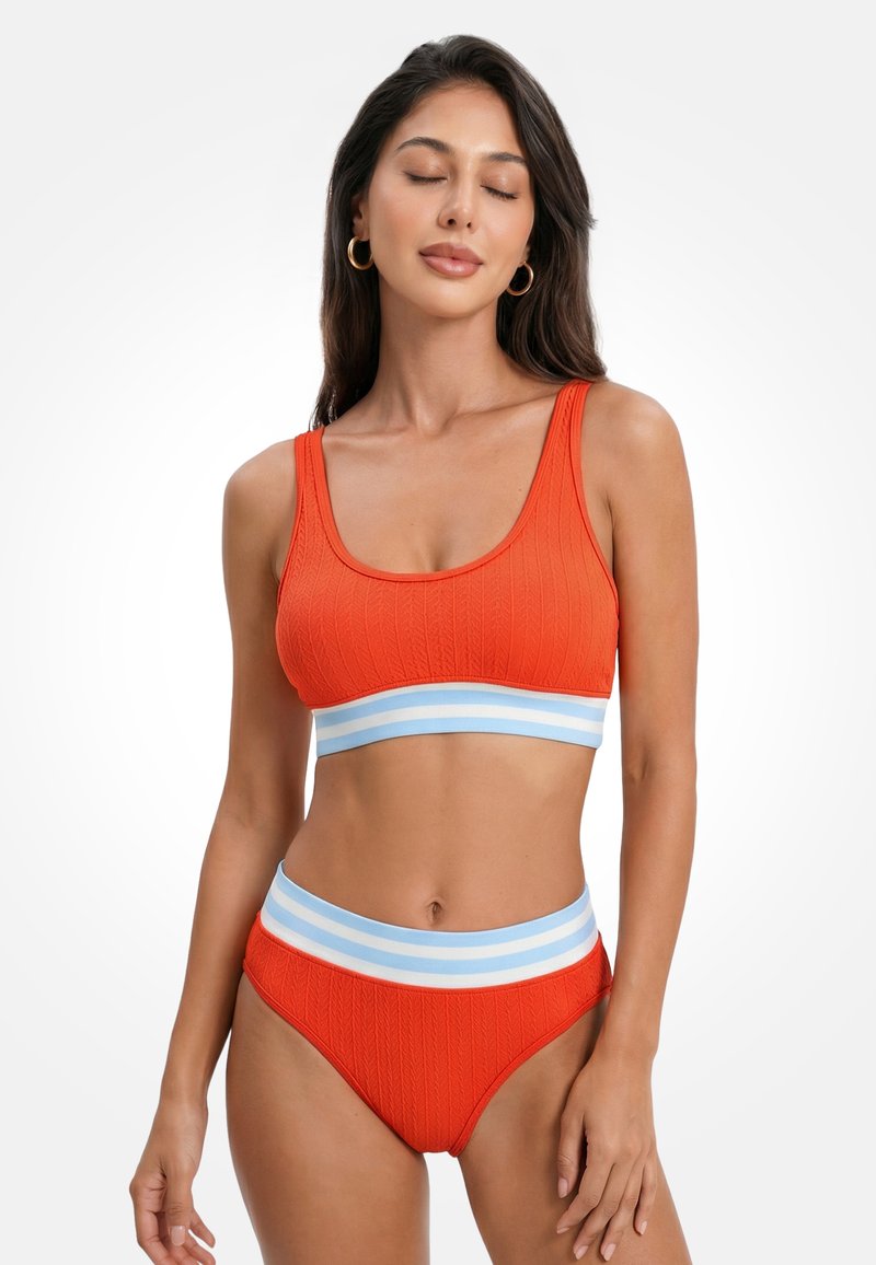 Vrouw met lang donker haar draagt oranje textuur zwemkleding in twee delen met lichtblauwe en witte gestreepte elastische banden, poserend met gesloten ogen.