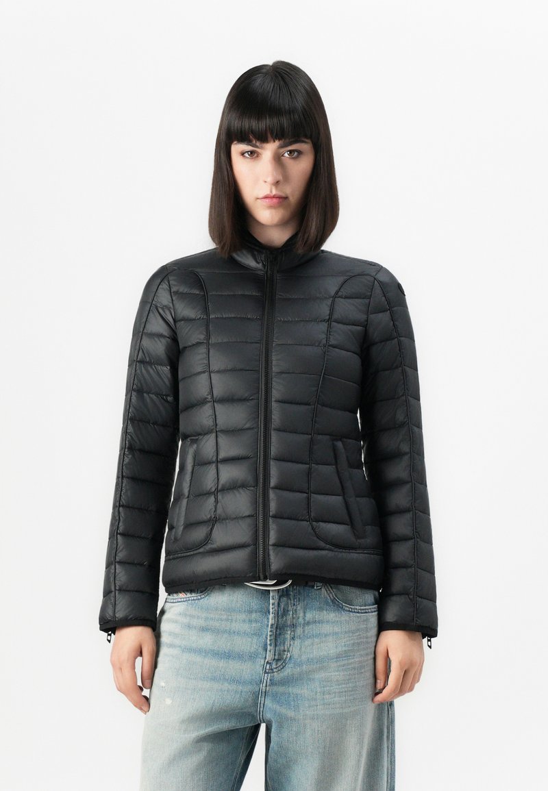 Giacca nera imbottita con colletto alto, cuciture orizzontali e zip frontale. Presenta tasche laterali e polsini regolabili. Indossata con denim chiaro.