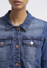 Jeansjacka i medelblått med knäppt front, två bröstfickor och klassisk krage. Lätt blekt struktur med bronsknappar.