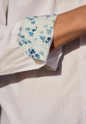 Hvid skjorte med mønstret manchet, der har blå blomsterdesigns på en lys baggrund. Glat stof med klassisk krave og knapdetalje.