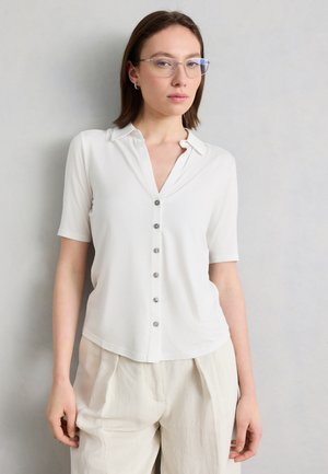 Νεαρή γυναίκα με κοντά καστανά μαλλιά, φορά καθαρές γυαλιά, λευκό κοντομάνικο πουκάμισο με κουμπιά και κρεμ παντελόνι μπροστά σε απλή επιτοίχια επιφάνεια.