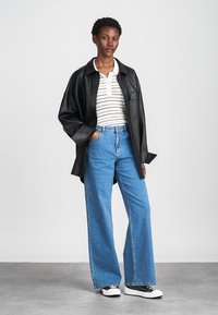 Musta nahkainen ylisuuria paita, raidallinen valkoinen polo, siniset leveälahkeiset farkut ja musta-valkoiset sneakersit. Suoraviivaiset tekstuurit ja yksinkertaiset kuviot.