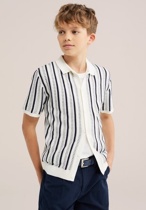 Fiú fehér és tengerészkék függőlegesen csíkos rövid ujjú kötött ingben, fehér póló fölött, tengerészkék nadrágban, zsebre tett kézzel, oldalra néz.
