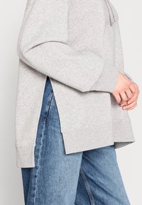 Person in einem hellgrauen langärmeligen Hoodie mit seitlichen Schlitzen und blauen Jeans, die Hände vor der Brust gefaltet.