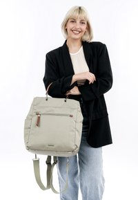 Grauer Stoffrucksack mit verstellbaren grünen Trägern, vorderer Reißverschlusstasche und braunen Lederelementen. Das Modell posiert in lässiger Kleidung.