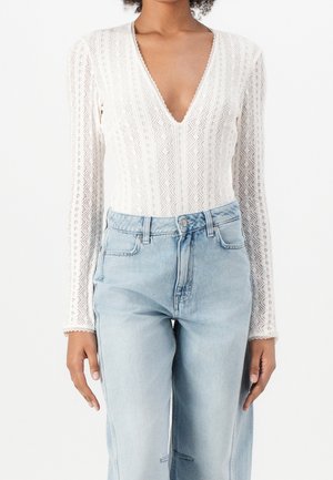 Kvinde iført hvid textureret, langærmet V-hals top stoppet ned i lyseblå, højtaljede jeans med forlommer.