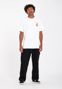 Hvid kortærmet t-shirt med farverig grafik på venstre bryst, parret med løse sorte cargo-bukser og sorte sko med hvide såler.