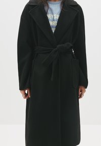 Čierny kabát vyrobený z mäkkej tkaniny, s opaskom v páse, veľkými klopami a bočnými vreckami. Textúrované dlhé rukávy, dĺžka po kolená.