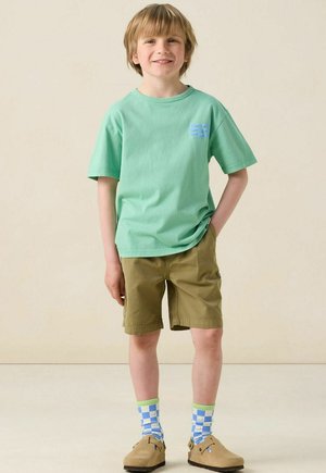 T-shirt en coton vert clair à manches courtes, shorts kaki, chaussettes fantaisie bleues et vertes, et chaussures slip-on tan.