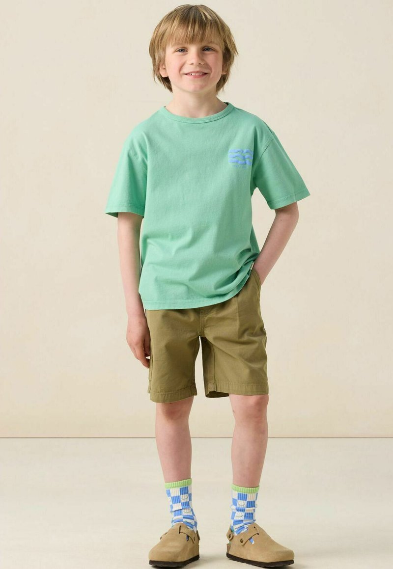 T-shirt en coton vert clair à manches courtes, shorts kaki, chaussettes fantaisie bleues et vertes, et chaussures slip-on tan.