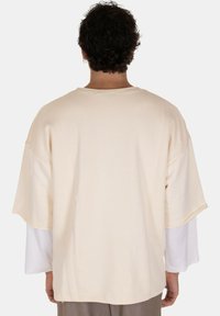 T-shirt beige oversize con maniche corte e scollo ampio. Il tessuto appare morbido. Indossato sopra una camicia a maniche lunghe bianca, visibile ai polsini.