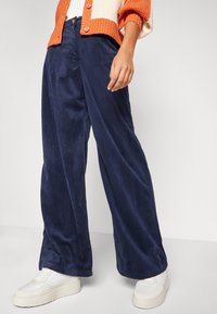 Široké kordové kalhoty v námořnické modři, s vertikálními žebry, předním zapínáním na knoflík a hladkým povrchem, doplněné bílými teniskami.