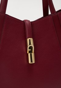 Hlboká červená kožená kabelka s textúrovaným povrchom a zlatým zapínaním pripevneným na popruhu blízko horného stredu.