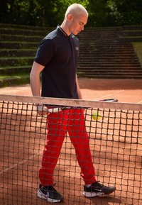 Navy polo shirt with decorative collar, red athletic pants, black sneakers. Player holding a tennis racket near a net on a clay court.