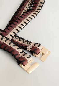 Cinturón de tela con secciones beige y marrón, costuras decorativas negras y cierre metálico dorado parcialmente abierto sobre un fondo claro.