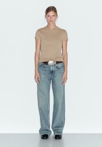 Beige aansluitend t-shirt met korte mouwen, gecombineerd met lichtblauwe baggy jeans en een donkerbruine riem met een grote zilveren gesp. Zwarte schoenen.
