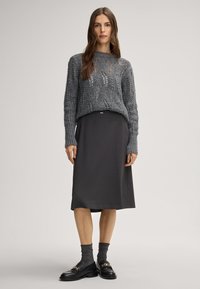 Maglione grigio a maglia con dettagli a punto aperto, abbinato a una gonna scura fino al ginocchio. La modella indossa scarpe nere e calze grigie texturizzate.
