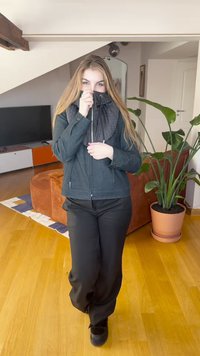 Tumehall lukuga jope tekstureeritud kraega, mustad püksid ja mustad kingad, taustaks puidust põrand ja toataimed.