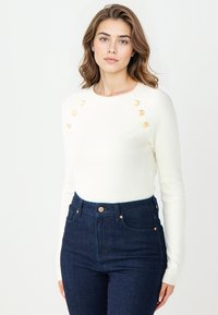 Pull blanc à côtes avec des boutons dorés aux épaules, associé à un jean bleu foncé taille haute. Texture douce, design ajusté.
