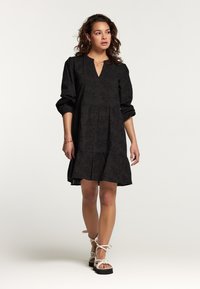 Rochie neagră cu decolteu în V, model floral texturat, mâneci lungi tip puff și fustă în straturi. Modelul poartă sandale cu platformă de culoare deschisă.