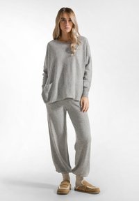 Maglietta grigia a maniche lunghe con tasca frontale abbinata a pantaloni larghi coordinati. Tessuto morbido e strutturato; completato con pantofole pelose beige e calze.
