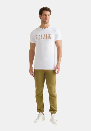 T-shirt bianco a maniche corte con "T'CLASSIE" stampato in lettere marroni testurizzate. Abbinato a pantaloni jogger verde oliva e sneakers bianche.