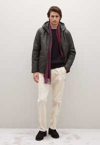 Giacca con cappuccio verde oliva, maglione a trecce blu navy, sciarpa a quadri rossa e blu, pantaloni crema e scarpe marrone scuro. Design e texture semplici.