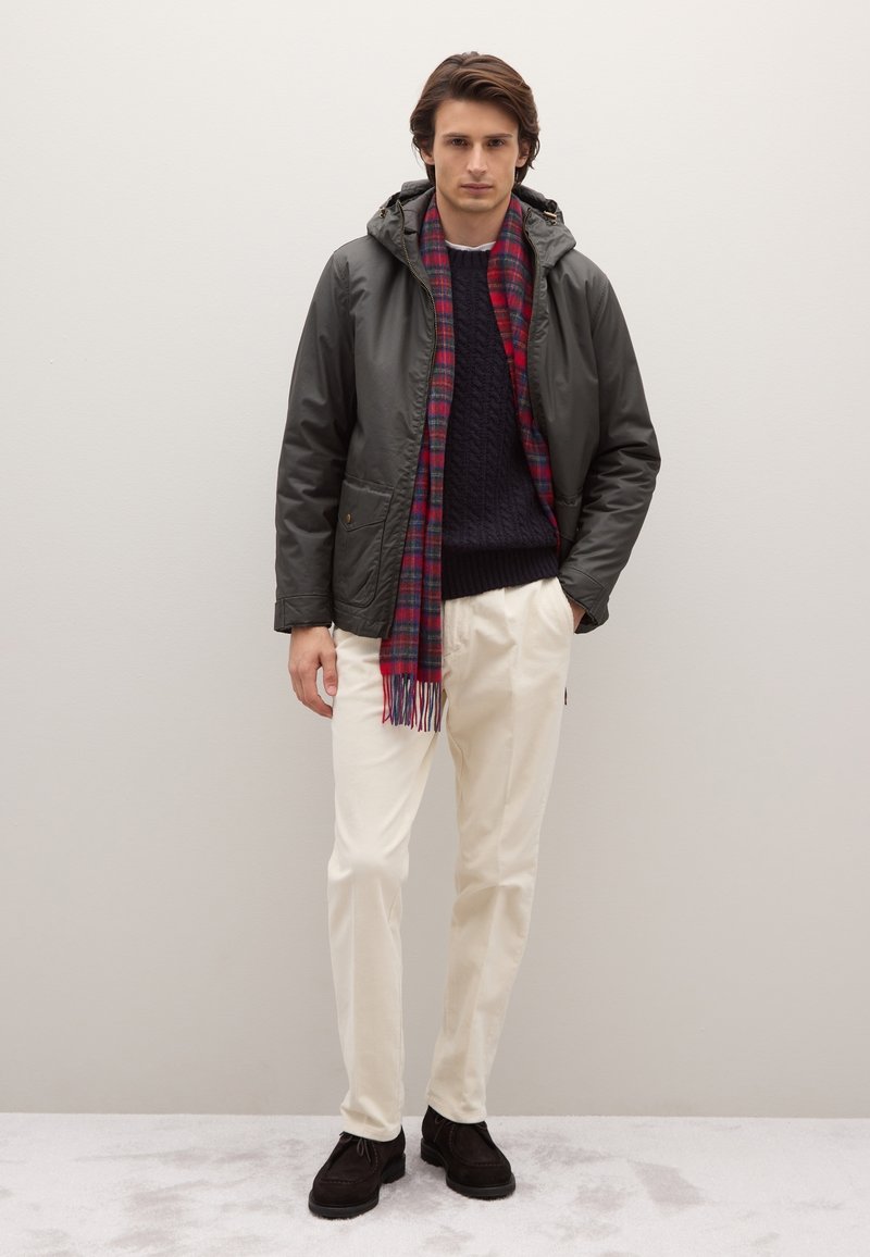 Giacca con cappuccio verde oliva, maglione a trecce blu navy, sciarpa a quadri rossa e blu, pantaloni crema e scarpe marrone scuro. Design e texture semplici.