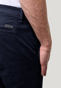 Marineblaue Hose mit glatter Textur, ausgestattet mit einer Seitentasche und einem Metalletikett, das an dem Bund angenäht ist. Hand auf der Hüfte abgestützt.