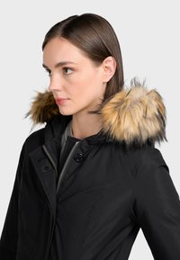 Čierna parka bunda s veľkým kožušinovým kapucňom, ktorý má hnedé a čierne zakončenia. Obsahuje zips a gombíkový uzáver a je elegantného dizajnu.