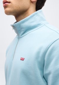 Jeune personne portant un sweat-shirt bleu clair à fermeture éclair quart avec un logo rouge Levi's sur la poitrine, sur un fond uni.
