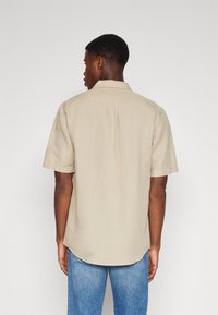 Chemise beige à manches courtes en tissu léger avec une coupe décontractée, dotée d'un empiècement au dos et d'un ourlet arrondi. Assortie avec un jean bleu.