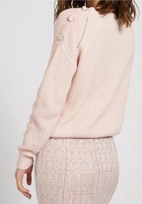 Pull rose clair avec une texture côtelée, orné de boutons argentés sur l'épaule et une coupe décontractée. Porté avec une jupe rose texturée.