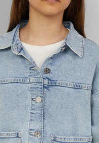 Helle blaue Jeansjacke mit Kragen, zwei vorderen Taschen, metallischen Knöpfen und subtilen Stickdetails. Hemd in schlichtem Weiß ist sichtbar.
