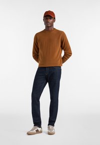 Maglione marrone, jeans blu scuro e sneakers color tan. Il cappellino è arancione. I materiali sembrano soffici e con una vestibilità casual, enfatizzando il comfort e la versatilità.