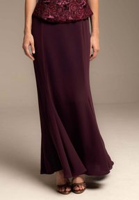 Una falda maxi de gasa burdeos, fluida, con una textura suave y un brillo sutil, combinada con sandalias marrones de tiras.