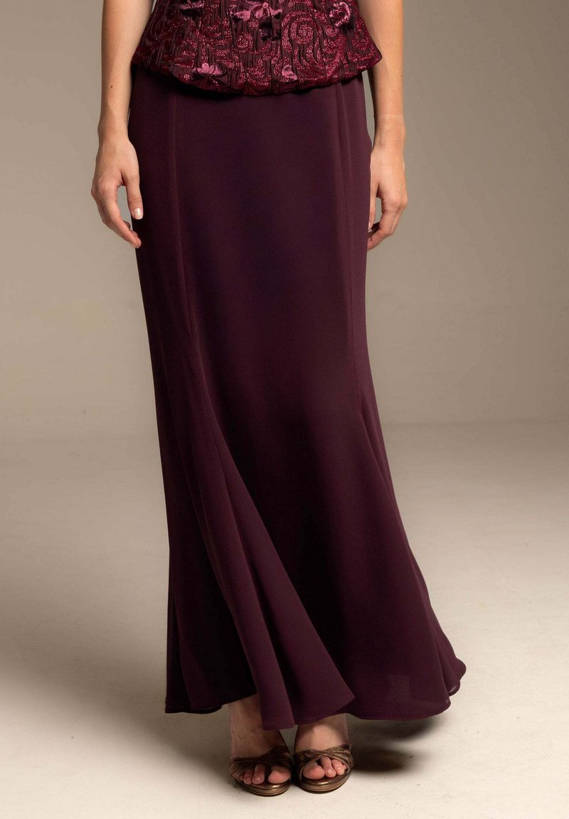 Una falda maxi de gasa burdeos, fluida, con una textura suave y un brillo sutil, combinada con sandalias marrones de tiras.