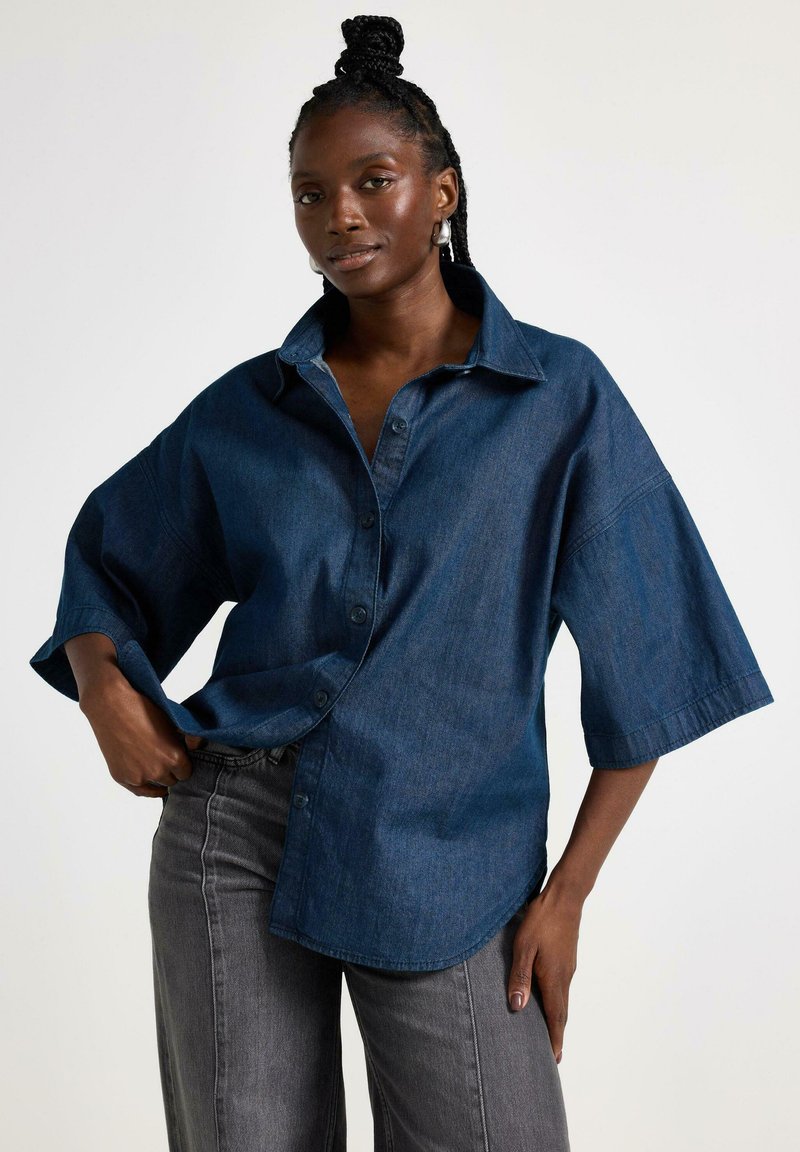 Blå denim oversized skjorte med knaplukning foran, krave og korte, brede ærmer. Kombineret med grå jeans. Glat tekstur.