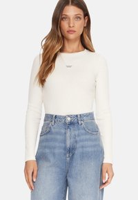 Maglietta bianca aderente a maniche lunghe con il logo "GUESS JEANS", abbinata a jeans di denim blu a vita alta, dalla vestibilità larga e con texture sbiadita.