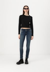 Black cropped long-sleeve top with side accents, paired with blue skinny jeans and chunky black ankle boots. Model wearing dark sunglasses.