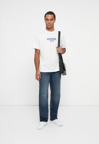 Hombre con camiseta blanca de Guess, jeans azules, zapatillas blancas y llevando una bolsa de hombro negra frente a un fondo blanco liso.