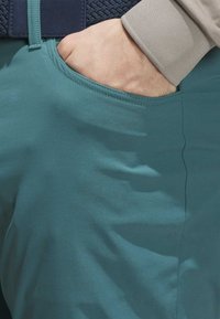 Türkise Hose mit glatter Textur und Seitentasche. Ein marineblauer, gewebter Gürtel setzt Akzente und wird durch einen hellen Sweatshirtbund kontrastiert.