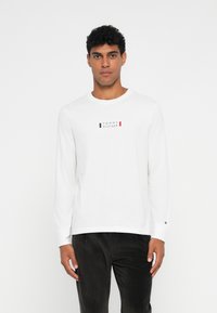 Bijela košulja dugih rukava s okruglim izrezom i malim logotipom "Tommy Hilfiger" u plavoj, crvenoj i crnoj boji na prsima. Pamuk.