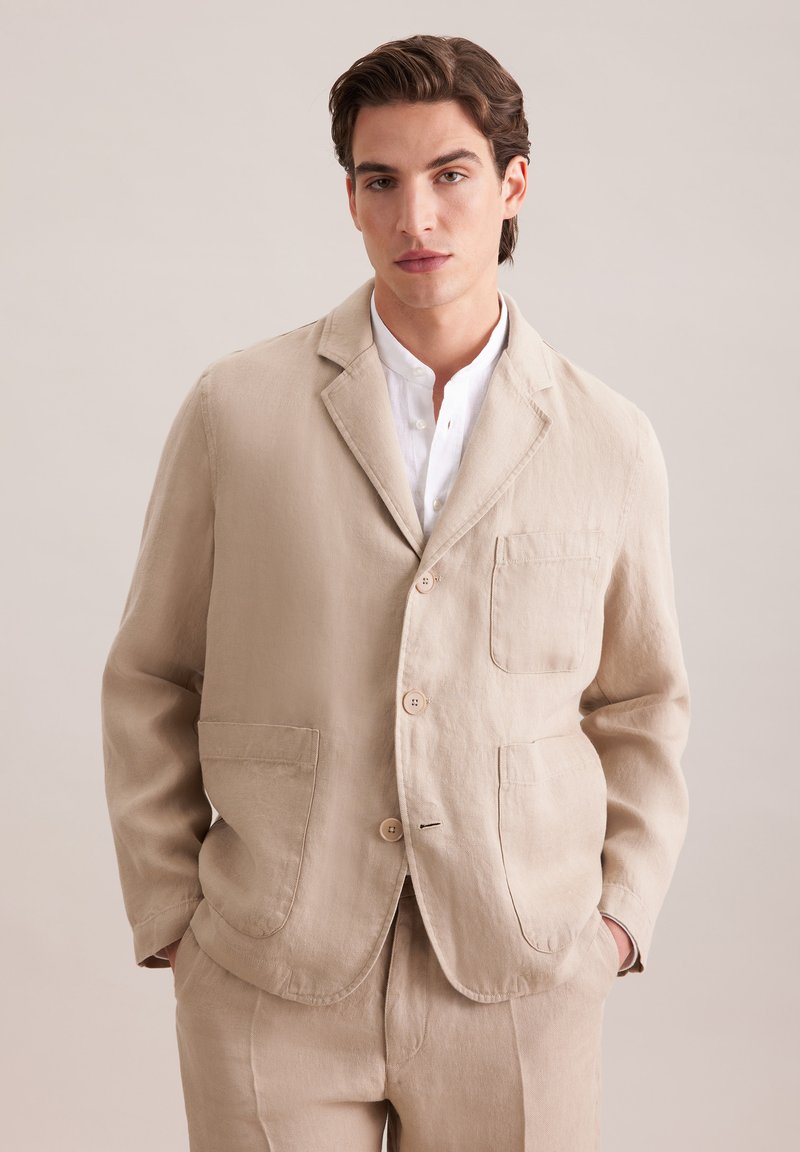 Jeune homme portant un costume en lin beige avec une veste à trois boutons et une chemise blanche, mains dans les poches, fond neutre.