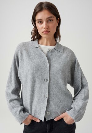 Giovane donna con capelli castani che indossa un cardigan grigio abbottonato sopra una maglia bianca, mani nelle tasche dei pantaloni neri, sfondo neutro.