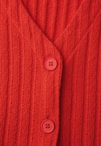 Roter Strickcardigan mit vertikaler Rippenstruktur und zwei runden roten Knöpfen, die vorne in der Mitte geschlossen sind.