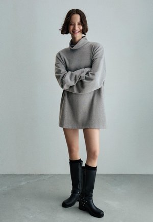 Grijze oversized sweater jurk met een hoge kraag en lange mouwen, gecombineerd met zwarte kniehoge laarzen met gesp-accenten. Soepele textuur.