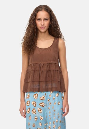 Giovane donna con capelli ricci che indossa un top marrone a strati senza maniche e pantaloni azzurri con motivi astratti marroni e bianchi.
